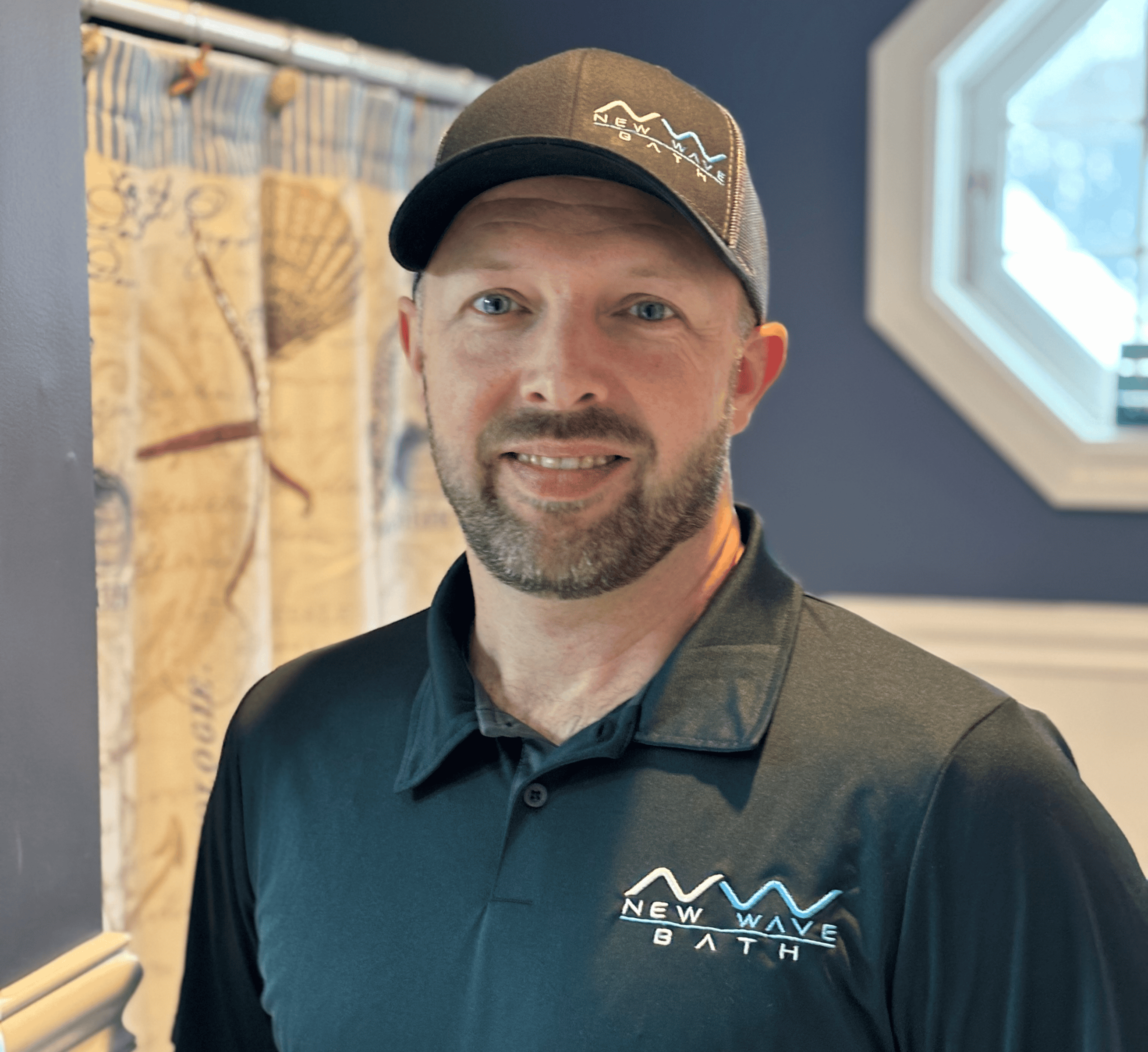 Modern Bathroom Installer Man in black "New Wave Bath" polo shirt and cap smiling indoors.