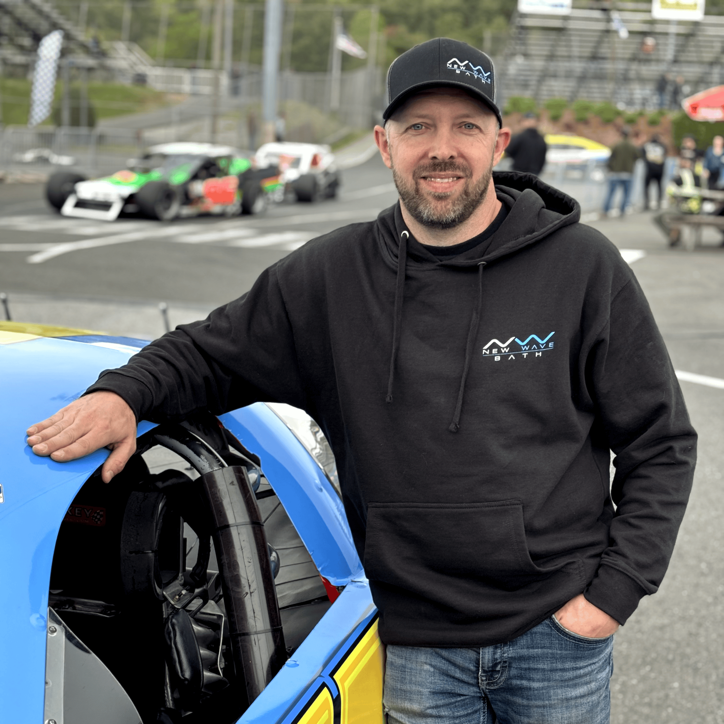 Man in black hoodie with New Wave Bath logo at a racing event.