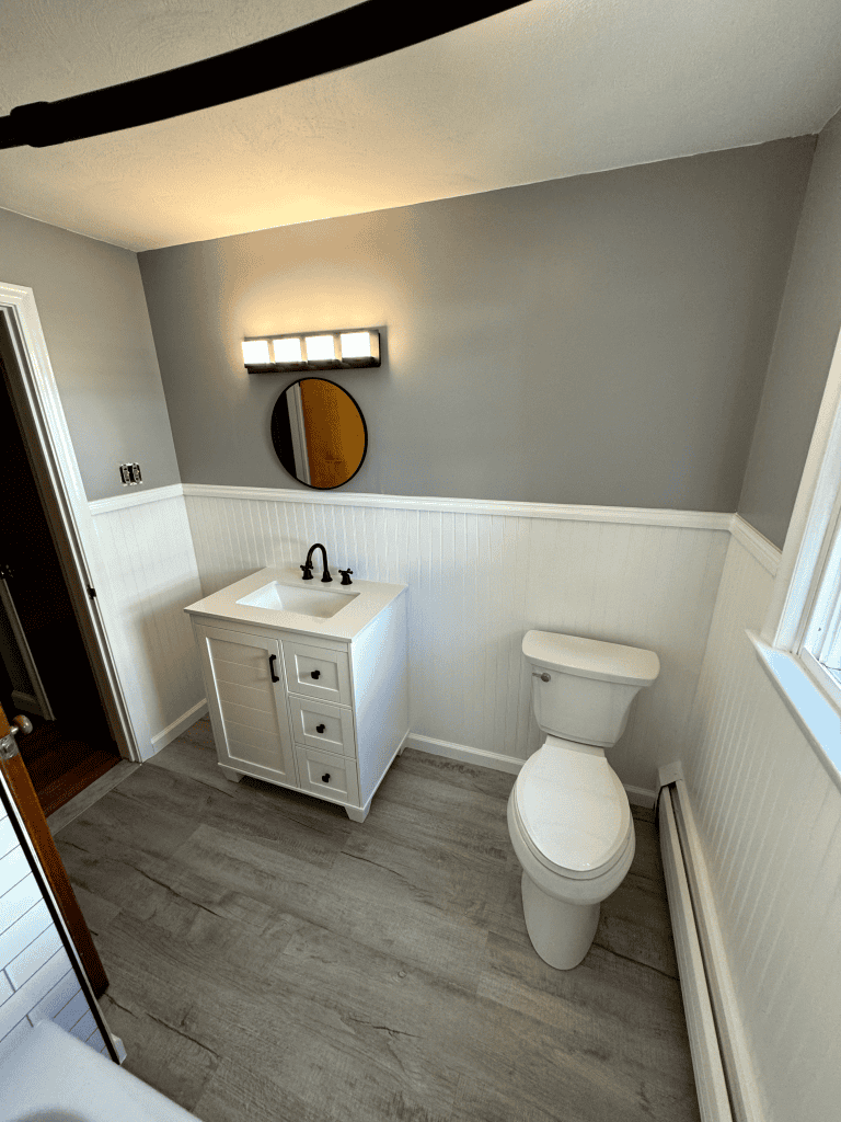 Small bathroom with white vanity, toilet, and gray walls, featuring a round mirror and natural light.