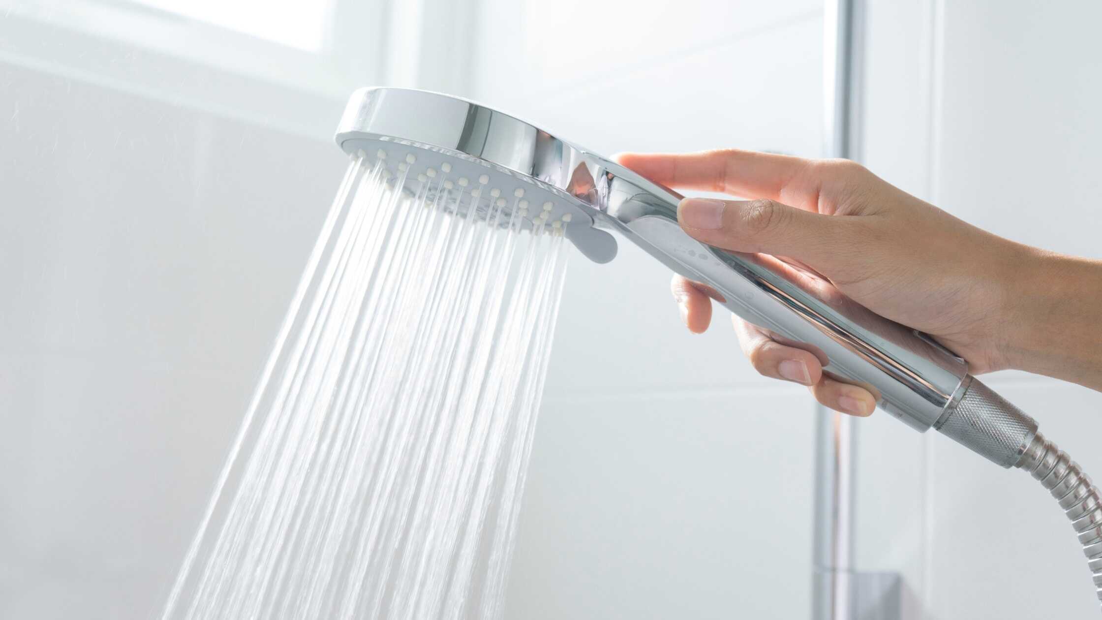 Hand holding a sleek, modern showerhead with water flowing in a bright bathroom.