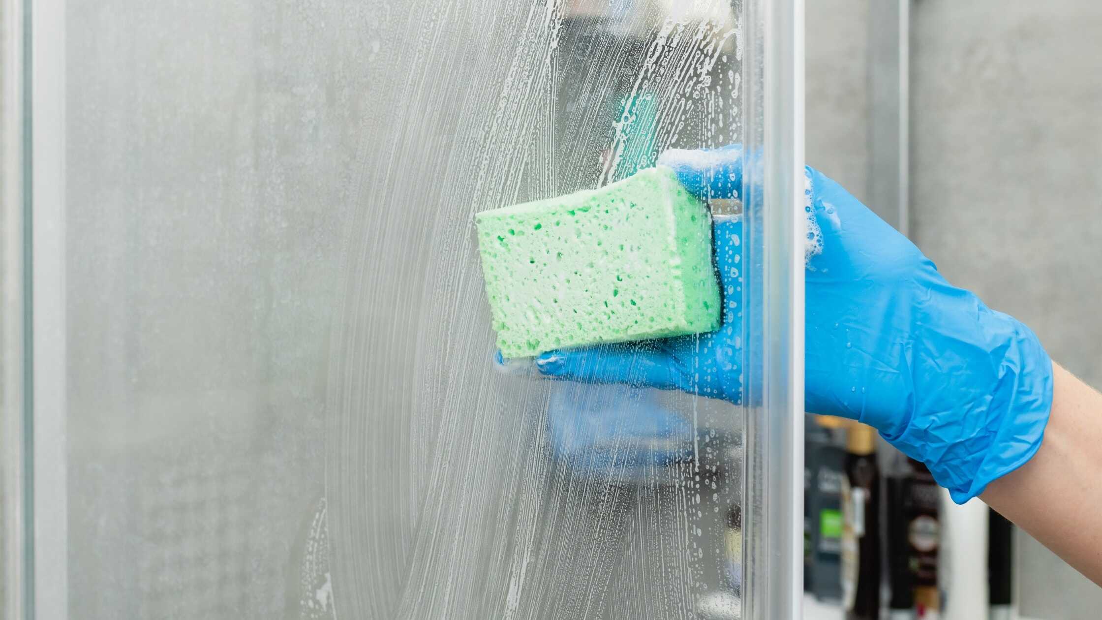 Hand in blue glove cleaning a glass shower door with a sponge and soap.