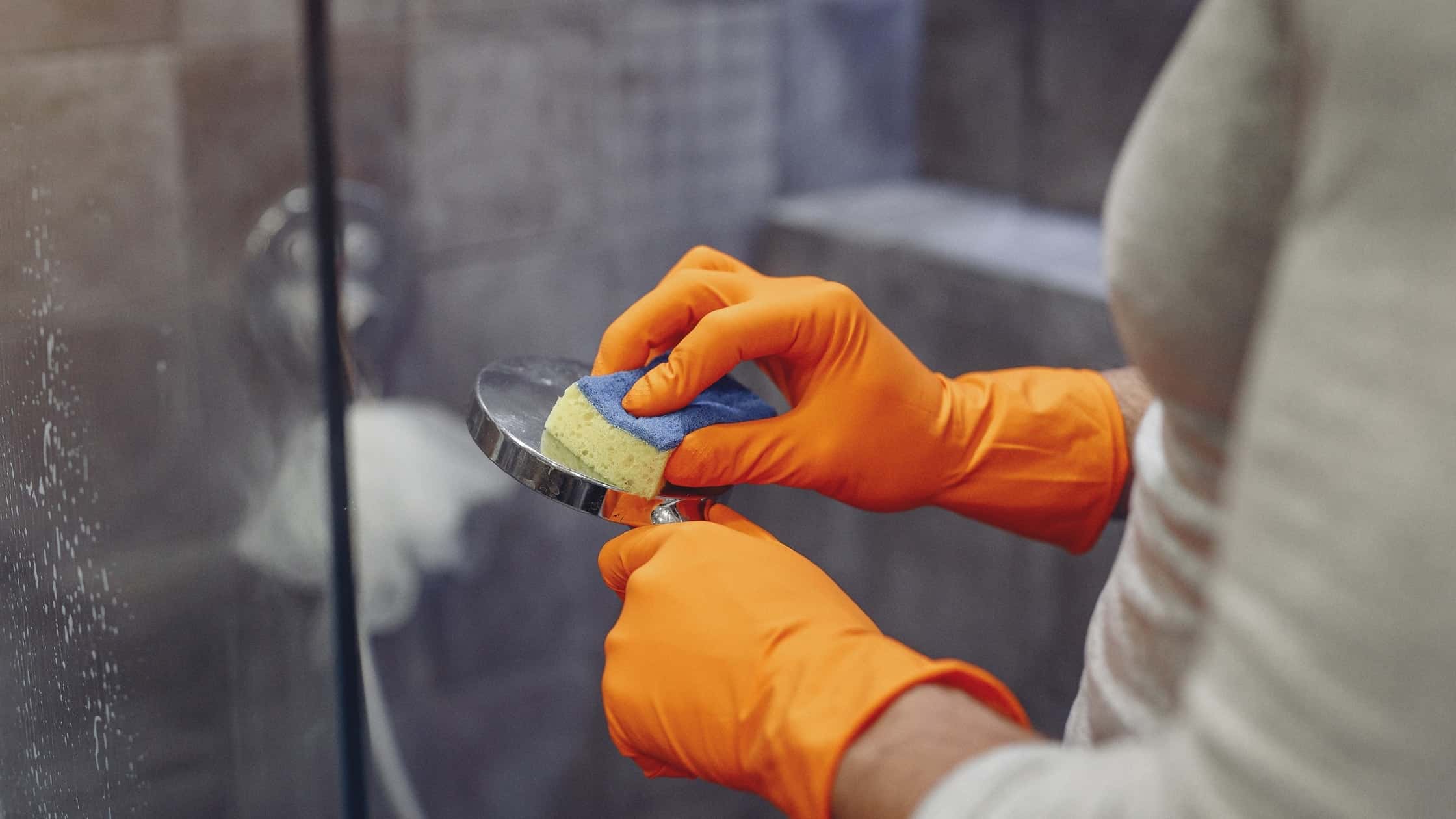 Person cleaning shower glass with sponge and orange gloves in bathroom.