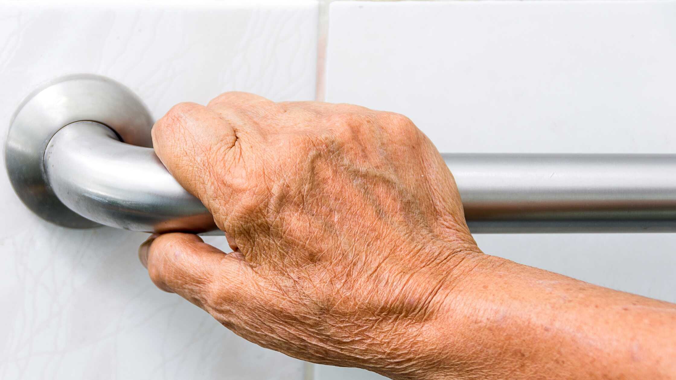 Close-up of an elderly hand gripping a bathroom safety handle for support.