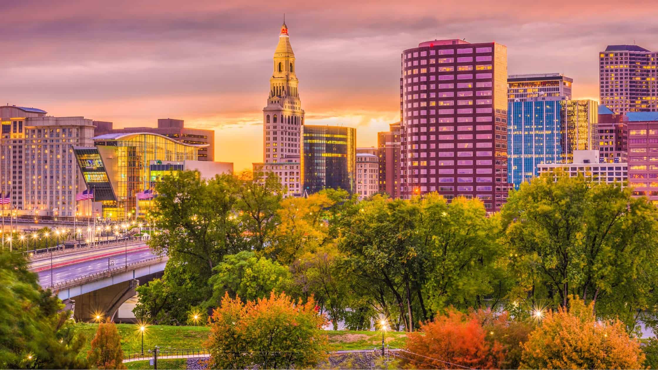 Cleveland city skyline at sunset with colorful sky and lush green trees in foreground.