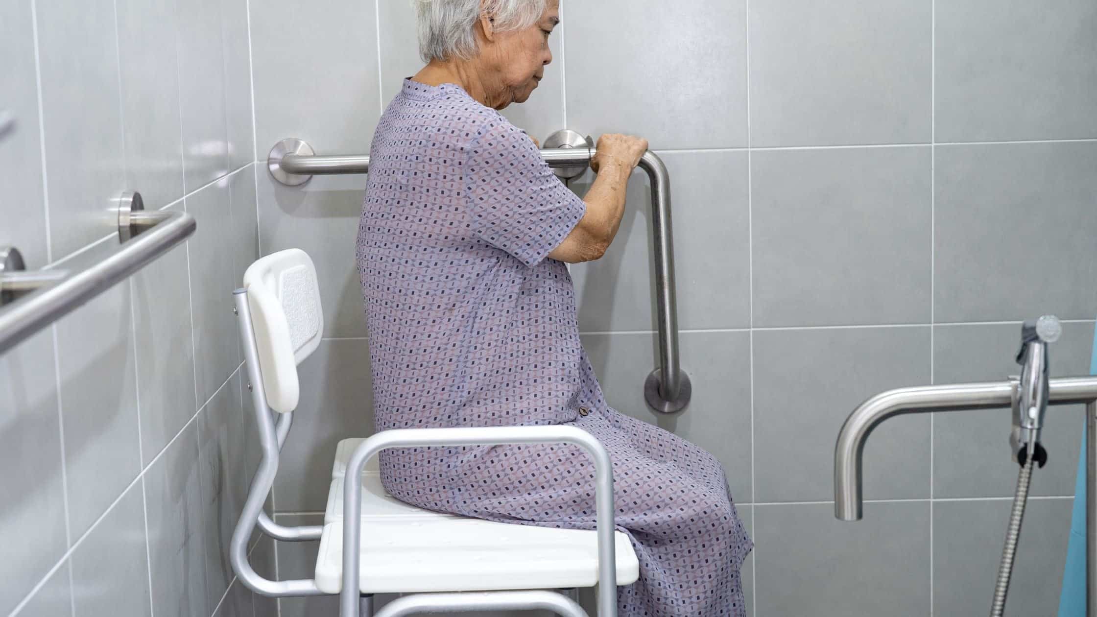 Elderly woman using a shower chair with grab bars in a bathroom for safe bathing.